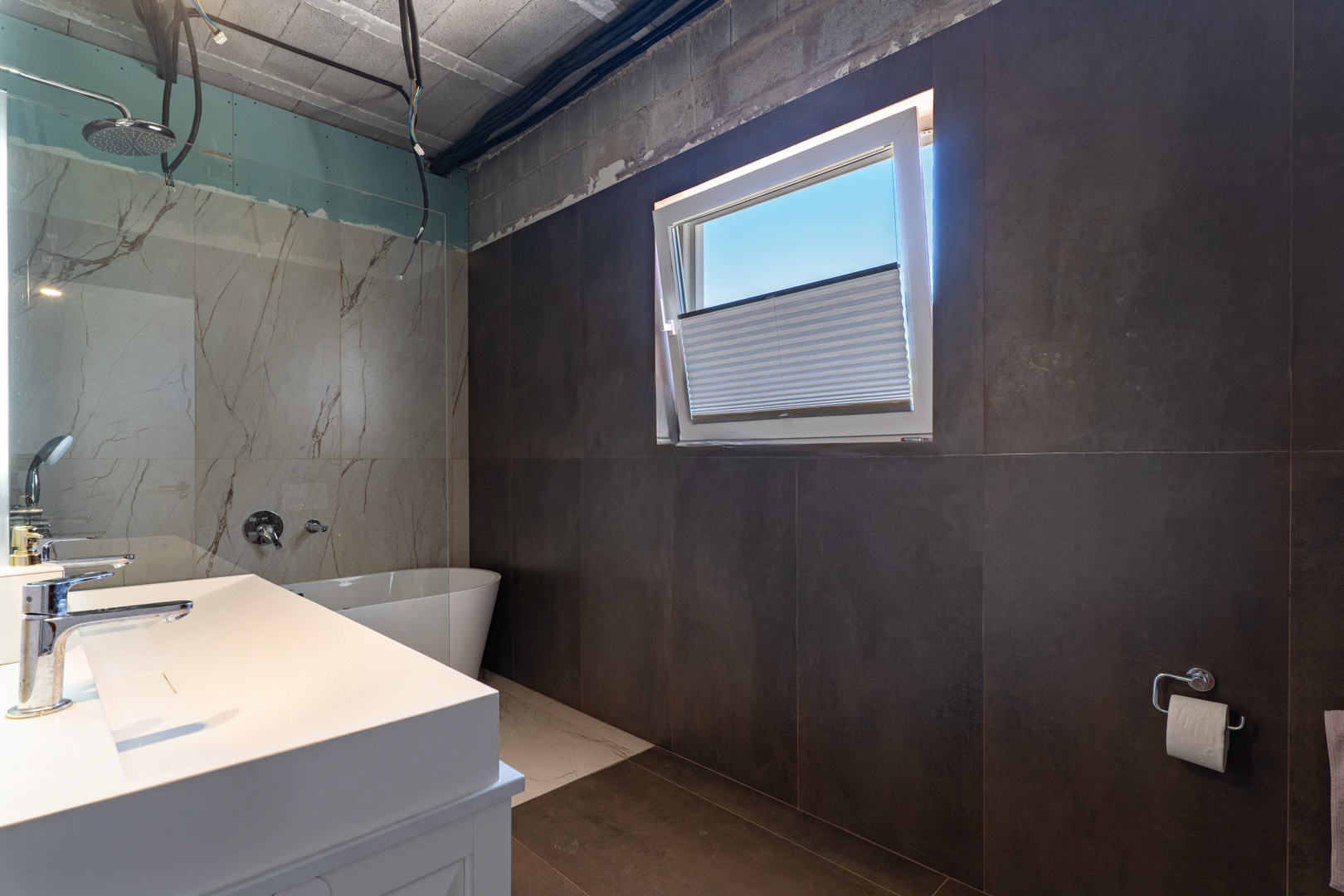 Badkamer in afbouw met dubbele wastafel, vrijstaand bad en inloopdouche met marmerlook. Grote donkere wandtegels combineren met het natuurlijke licht van het draaikiepraam.