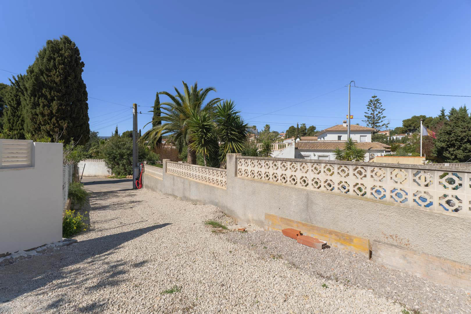 Buitenruimte van het perceel met een oprit van wit grind en een decoratieve betonnen omheiningsmuur, omgeven door mediterrane palmen en beplanting.