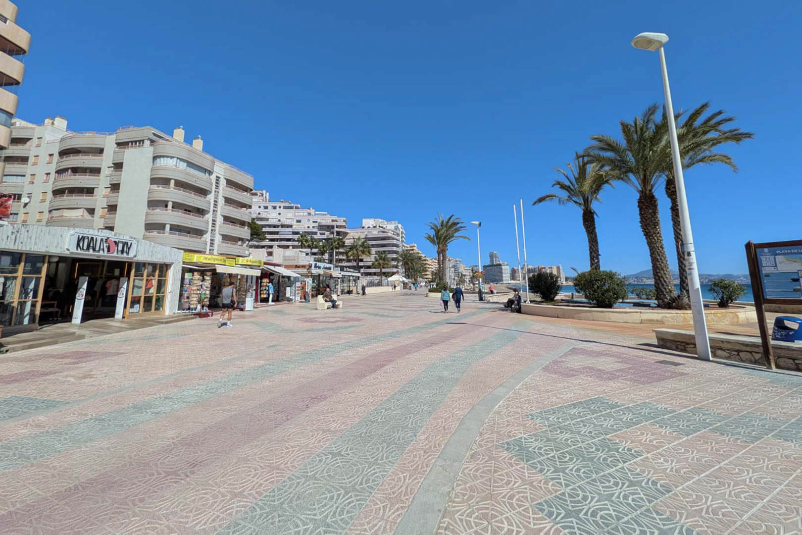 De boulevard van Calpe met decoratieve bestrating, omzoomd door lokale winkels, palmbomen en appartementencomplexen onder een heldere hemel.
