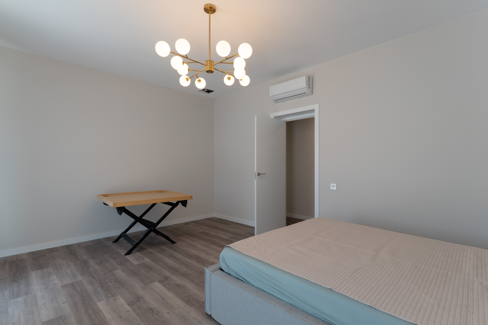 Eigentijdse slaapkamer met grijze laminaatvloer, neutrale wanden en een moderne gouden kroonluchter. Voorzien van airconditioning en een minimalistische witte deur.
