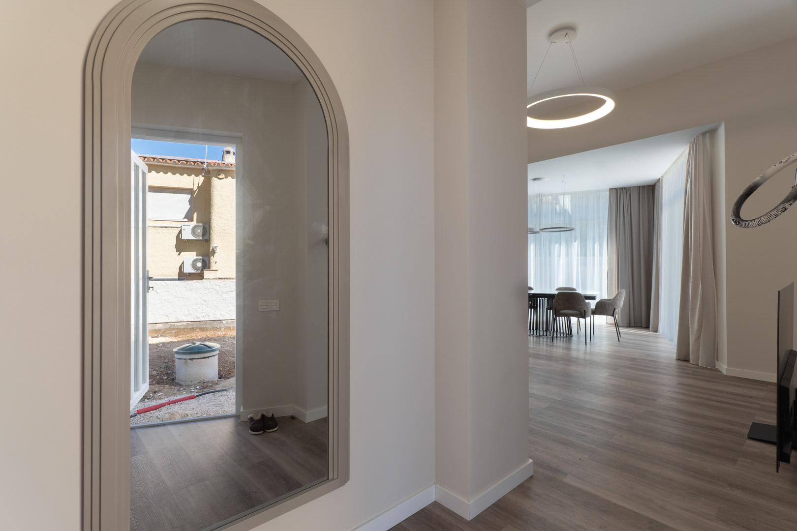 Entree met een boogspiegel die het exterieur reflecteert, leidend naar een minimalistische woonkamer met houtlook vloer en een ronde hanglamp.