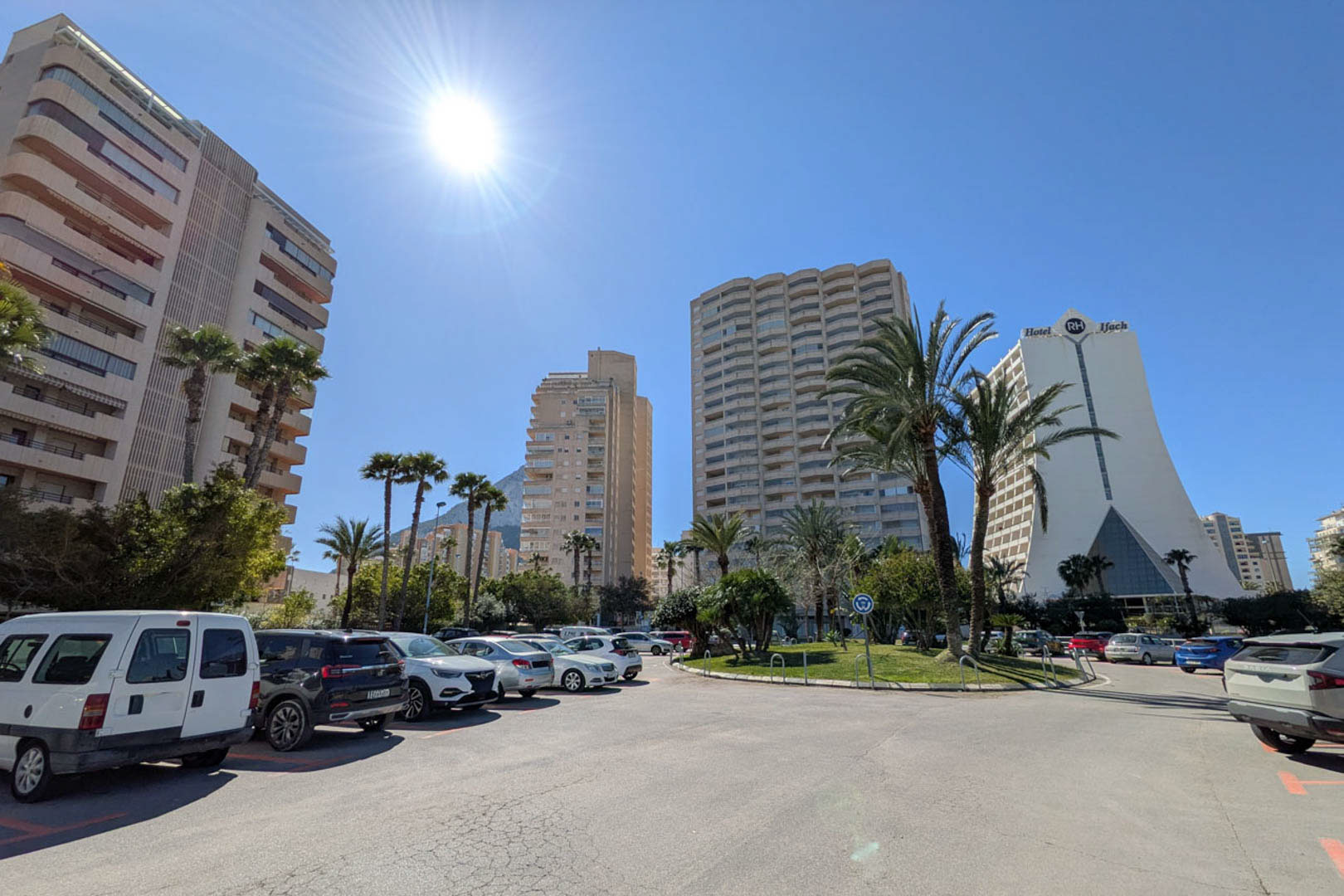 Exterieur van een stedelijke zone in Calpe, met hoge appartementsgebouwen, het RH Ifach Hotel en openbare parkeerplaatsen onder een strakblauwe lucht.