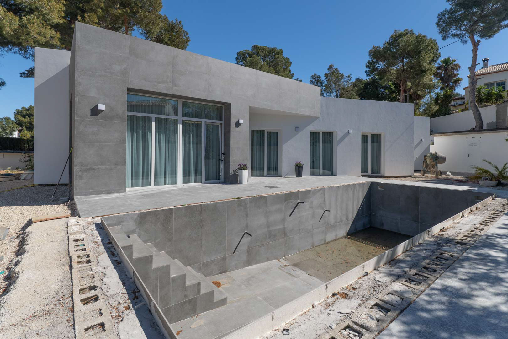 Gelijkvloerse minimalistische villa met grijze porseleinen gevelbekleding en een zwembad in aanbouw, omgeven door mediterrane pijnbomen.