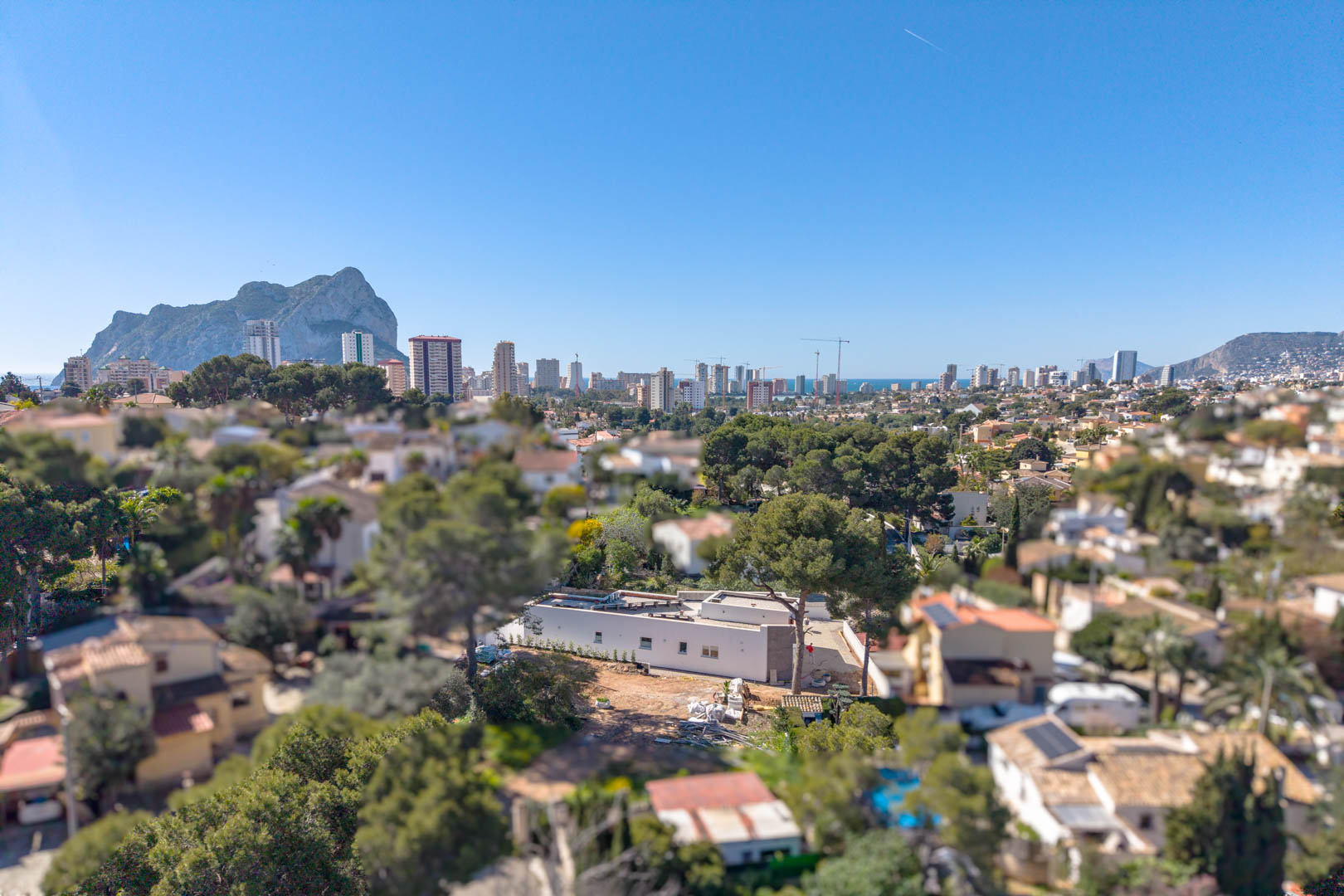 Luchtfoto van de minimalistische villa met de Peñón de Ifach op de achtergrond. De gelijkvloerse architectuur en het perceel in afbouwfase zijn duidelijk zichtbaar.