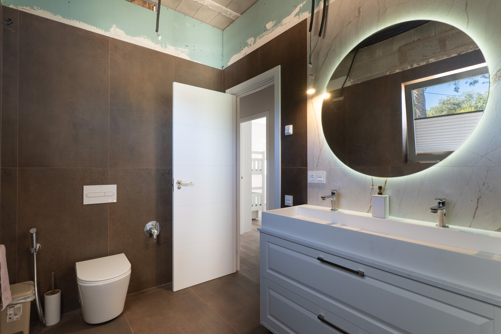 Moderne badkamer met dubbele wastafel, verlichte ronde spiegel en donkerbruine tegels. Voorzien van een hangend toilet en minimalistisch wit meubel.