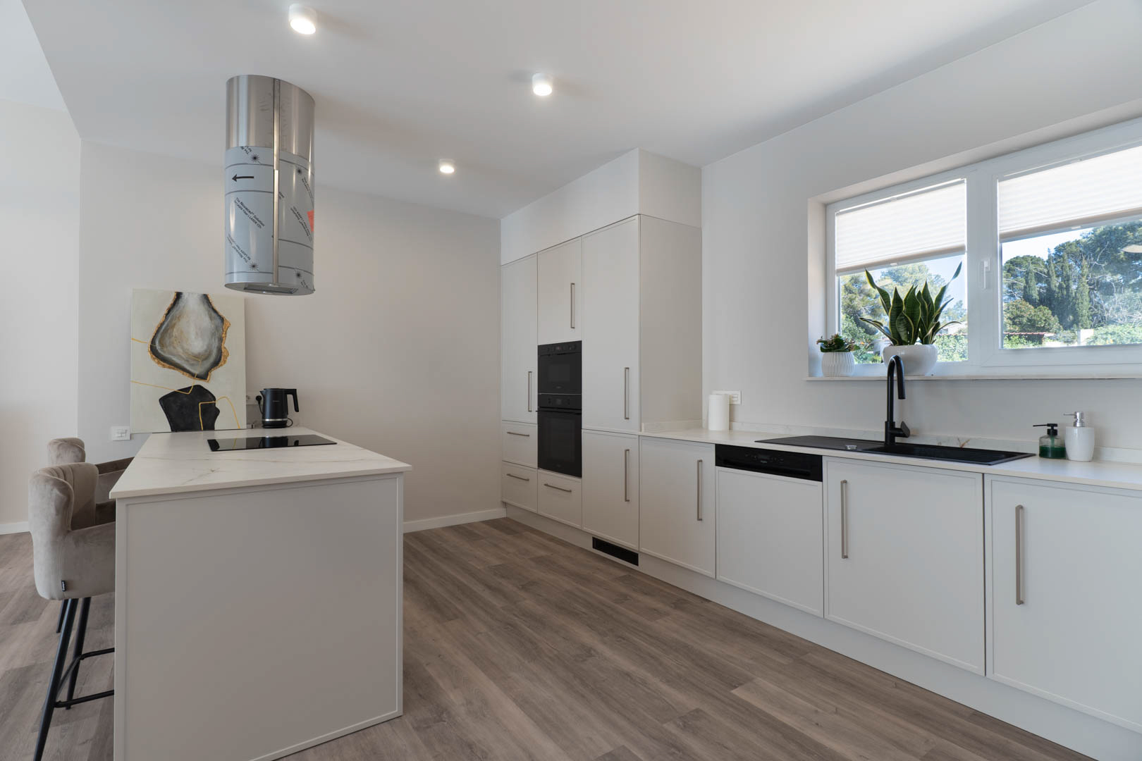 Moderne keuken met kookeiland, cilindrische afzuigkap en inbouwapparatuur. Witte kasten worden gecombineerd met zwarte accenten en een warme laminaatvloer met houtlook.