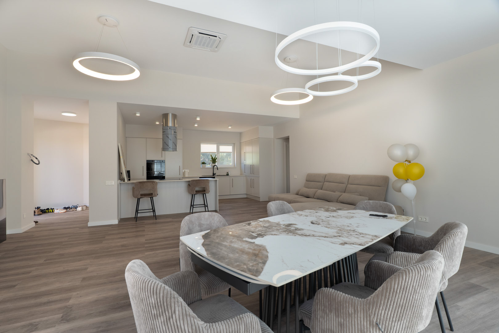 Open woonruimte met een marmeren eettafel, gestoffeerde stoelen en een minimalistische witte keuken met kookeiland en moderne ronde LED-verlichting.