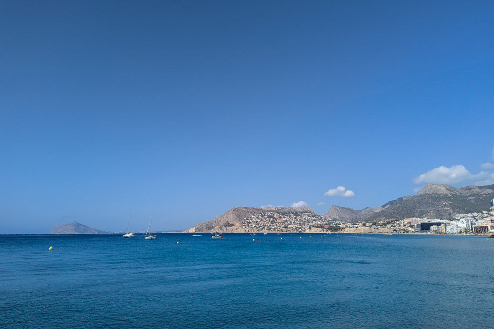 Panoramisch uitzicht op de Middellandse Zee voor de kust van Calpe, met zeilboten en de bergachtige kustlijn onder een helderblauwe hemel.