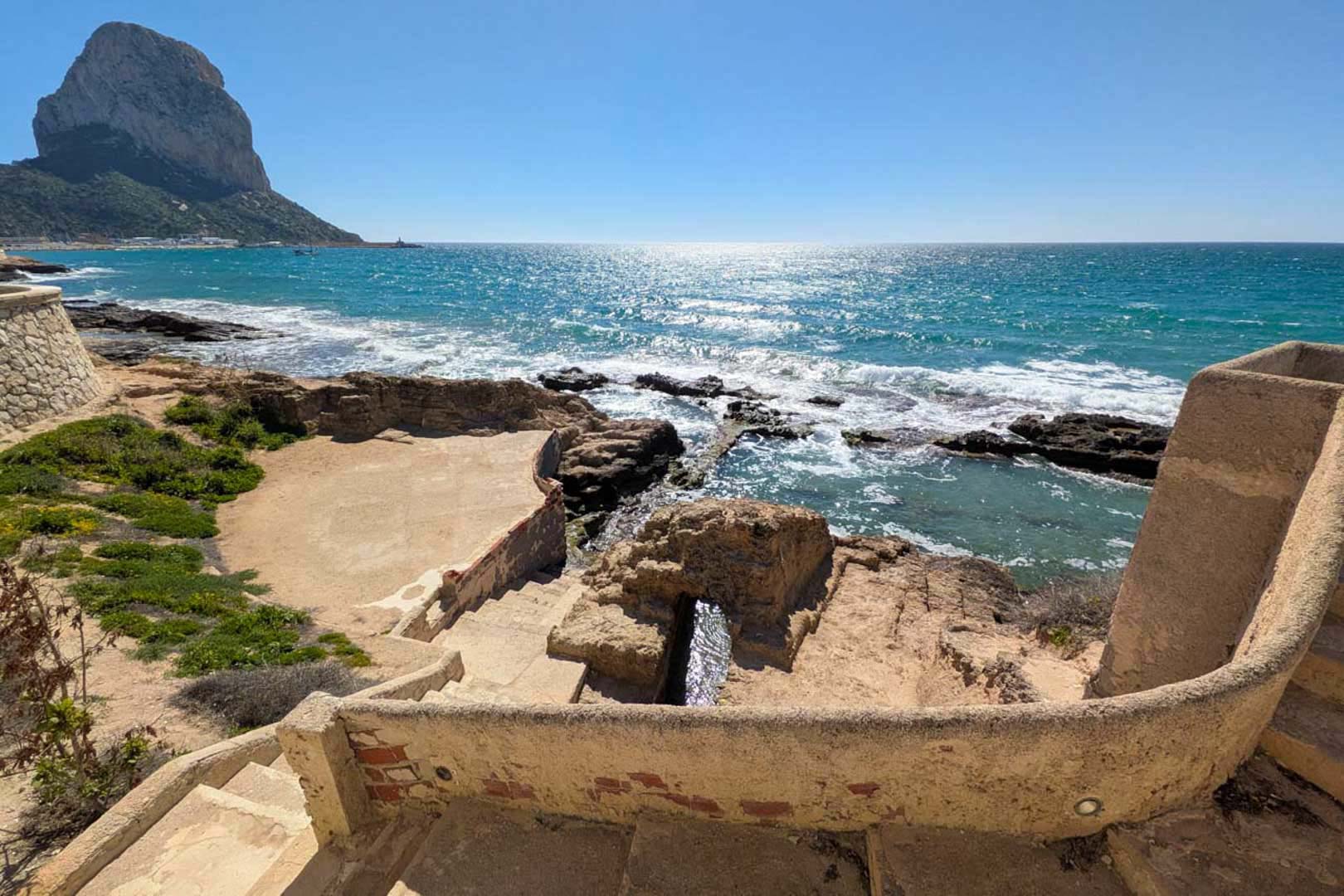 Panoramisch uitzicht op de Peñón de Ifach vanaf een stenen terras, met directe toegang tot een baai met turquoise water en de kustlijn.