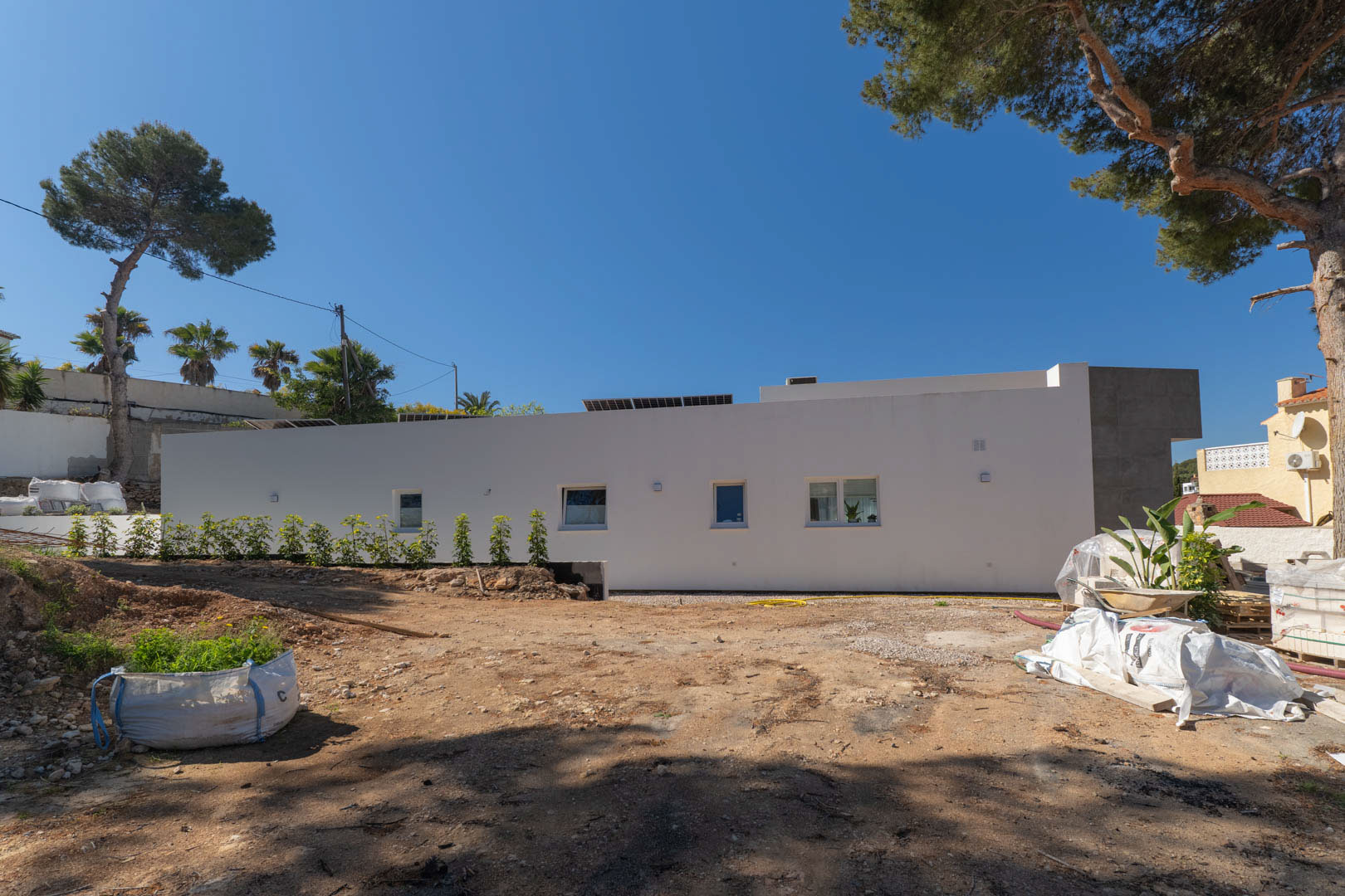 Zijgevel van de minimalistische villa met zonnepanelen, waarbij het buitenterrein in voorbereiding is en omringd wordt door volwassen pijnbomen.