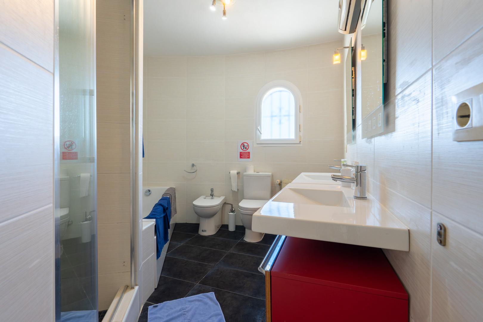 Badkamer met dubbele wastafel op een rood meubel, bidet, toilet en ligbad. Voorzien van lichte wandtegels, een donkere vloer en een klein boograam.