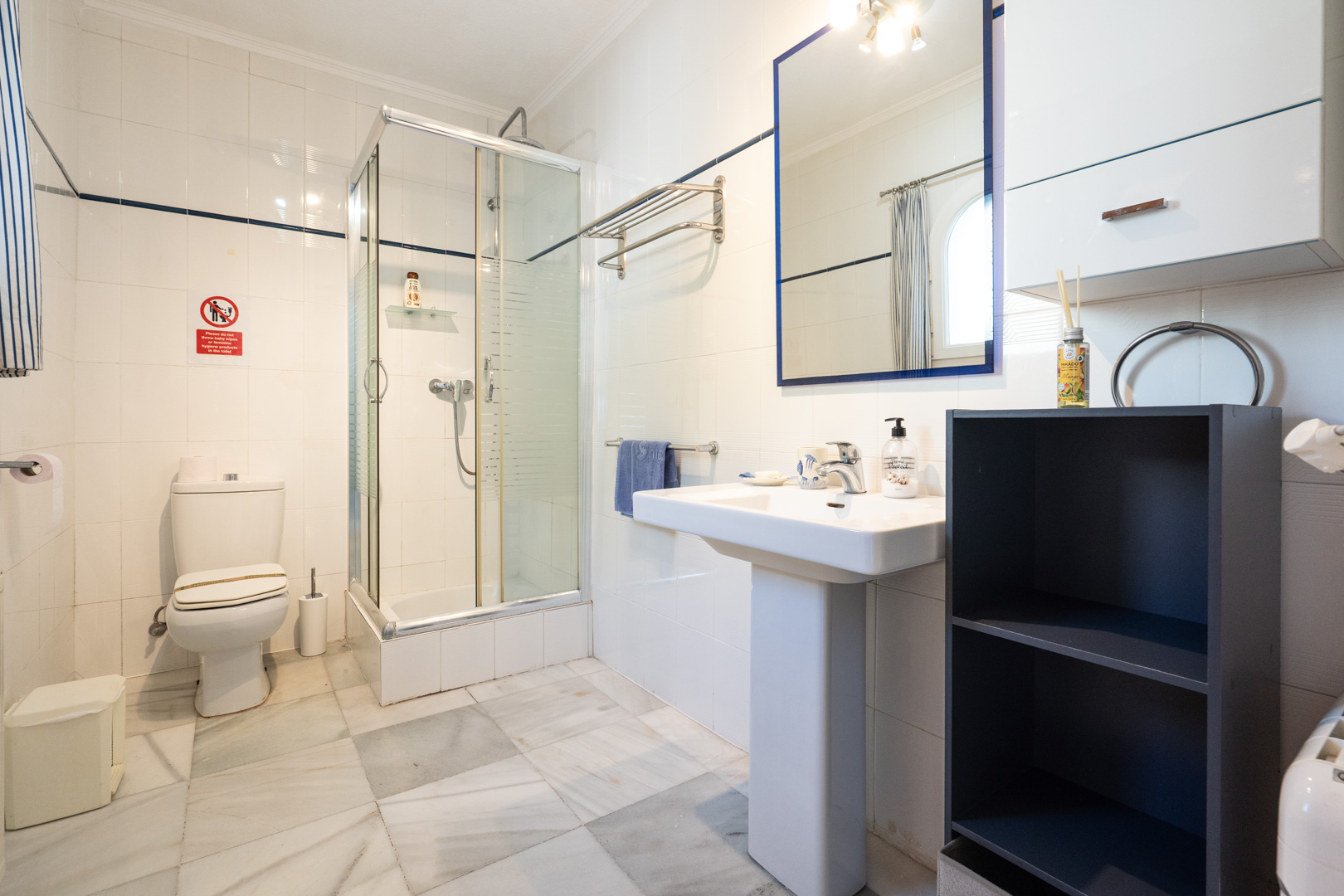 Badkamer met witte wandtegels, marmeren vloer, een glazen douchecabine en een kolomwastafel met een spiegel met blauwe lijst.