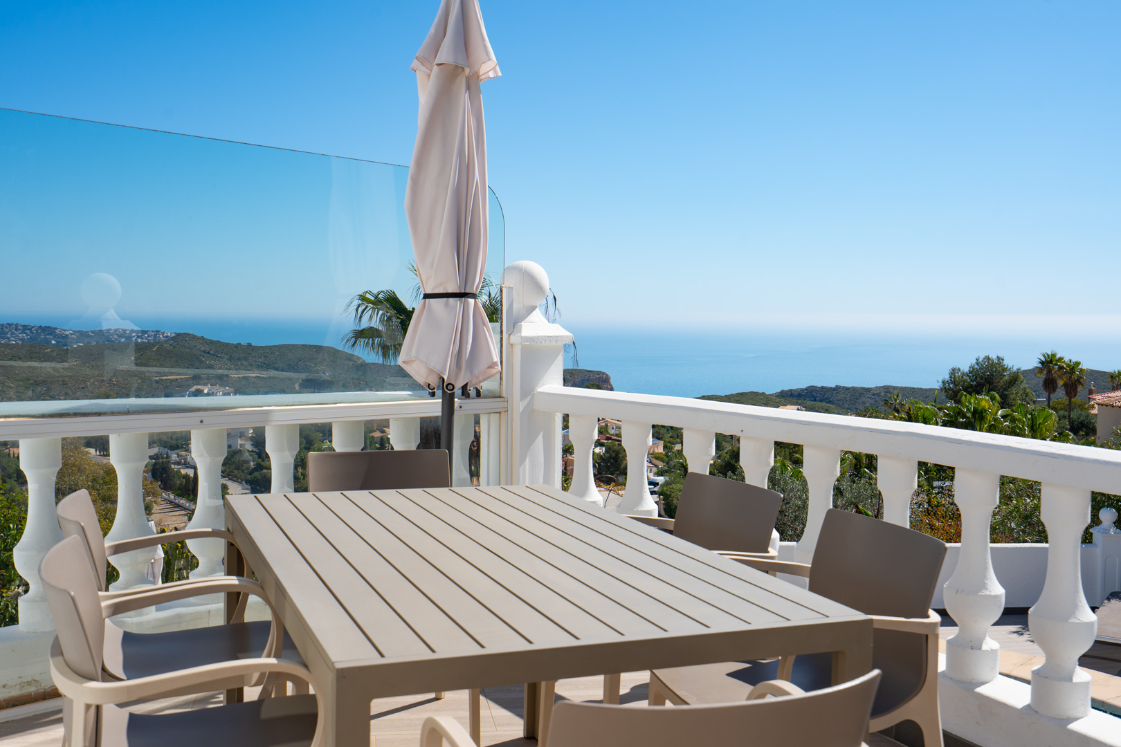 Buitenterras met eettafel en stoelen, afgezet met een witte balustrade en glazen panelen, met panoramisch uitzicht over de Middellandse Zee en de kustlijn.