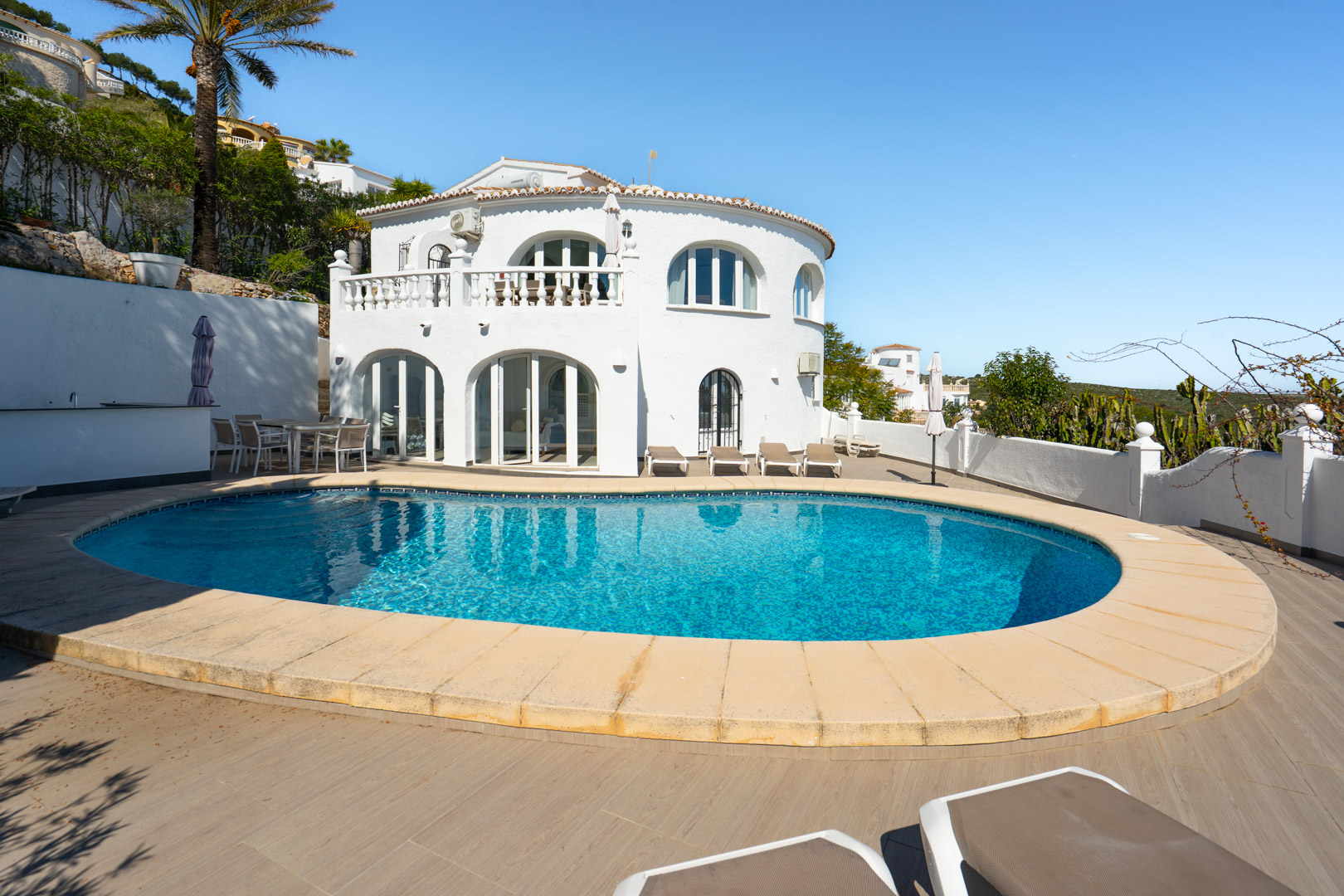 Exterieur van de villa met een niervormig zwembad, ruim terras met houtlook tegels en een witte gevel met boogvensters en een klassieke balustrade.