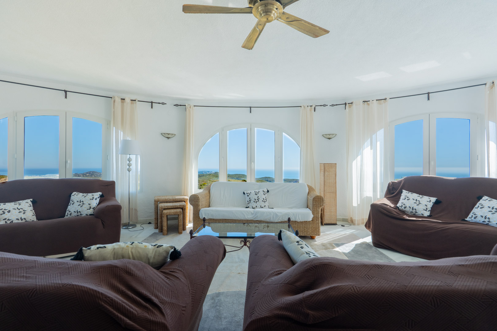 Ronde woonkamer met boogvensters die een panoramisch uitzicht op zee bieden. De ruimte is voorzien van een plafondventilator, rieten meubels en veel natuurlijk licht.