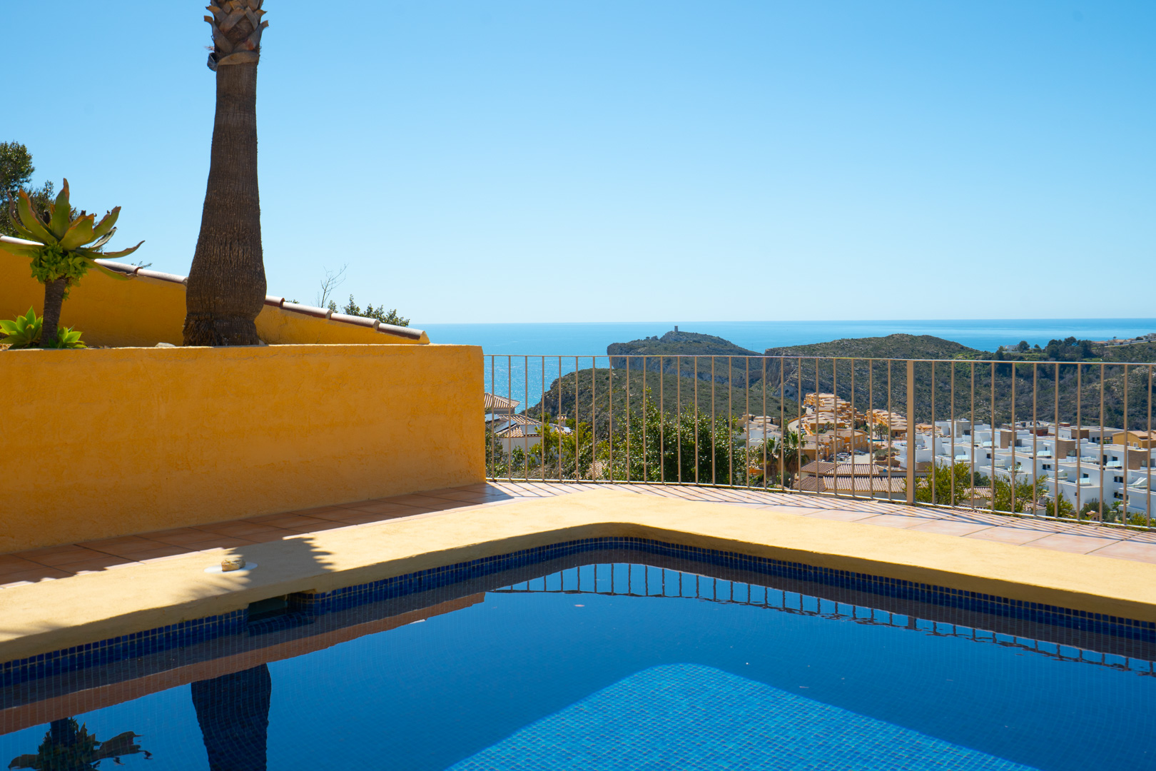 Blauw betegeld zwembad en zonneterras met panoramisch uitzicht op de Middellandse Zee en de landtong Cap d'Or in Moraira, omheind met een metalen reling.