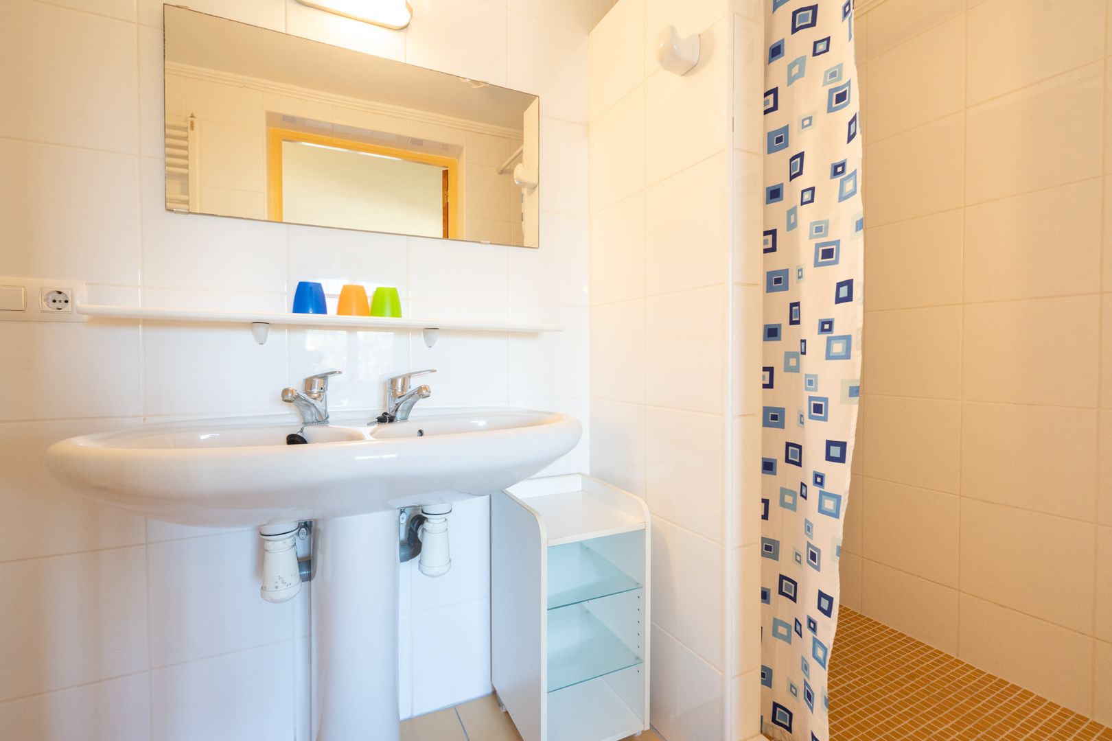 Functionele badkamer met een dubbele wastafel op zuil, witte wandtegels en een doucheruimte met patroongordijn en okerkleurige vloertegels.