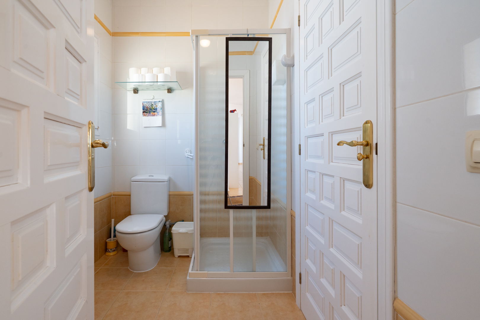 Functionele badkamer met wit-beige betegeling, voorzien van een toilet en douchecabine met spiegel, bereikbaar via witte paneeldeuren.