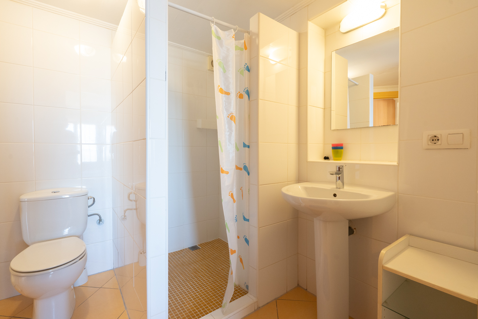Functionele badkamer met witte wandtegels, inloopdouche met gordijn, kolomwastafel en klassiek sanitair in lichte tinten.