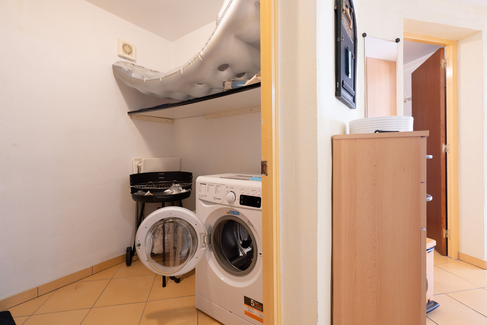 Functionele wasruimte met een voorlader wasmachine, opbergplank en lichte tegelvloer, grenzend aan de overige woonvertrekken.