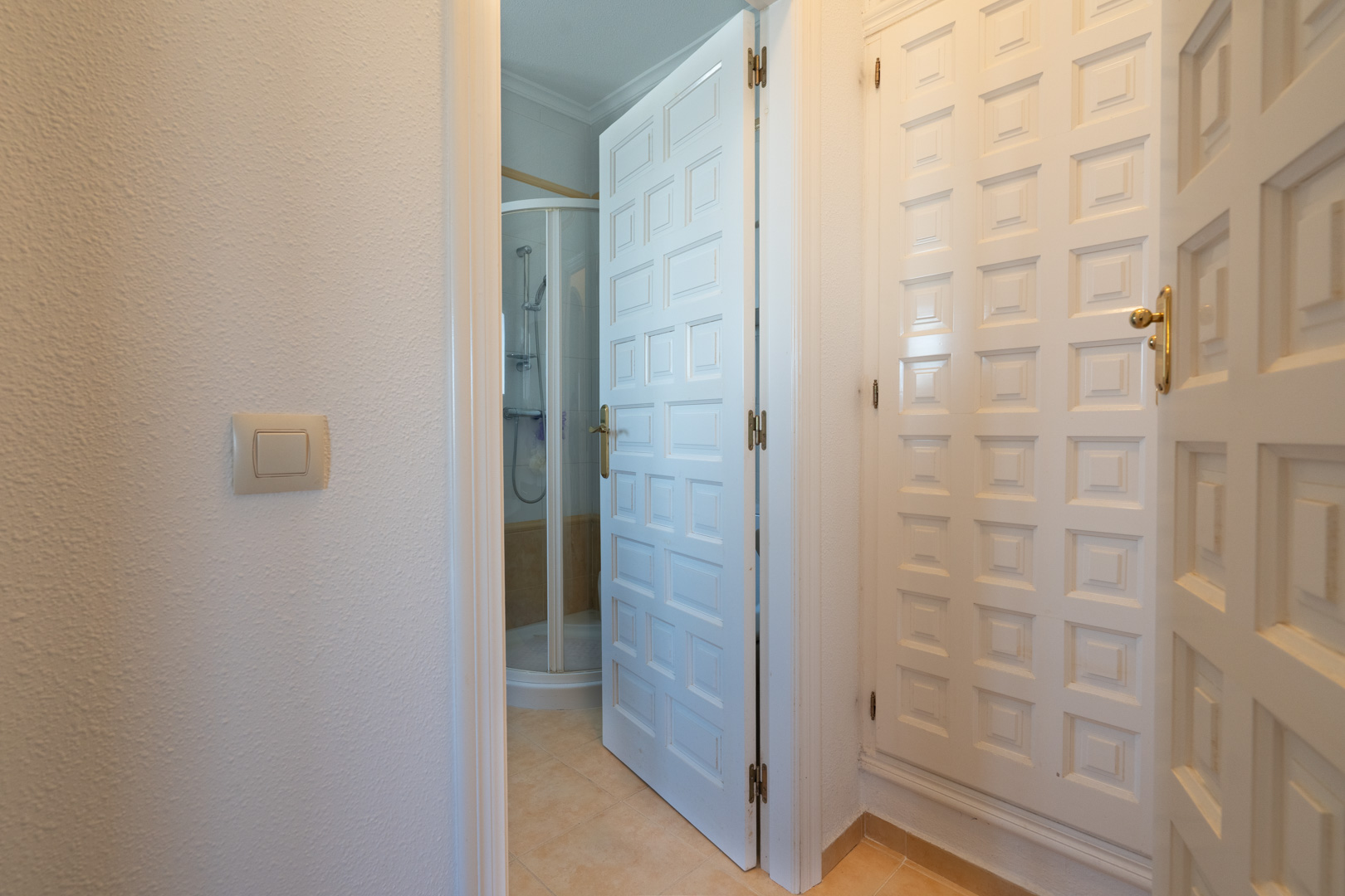 Gang met wit gepaneelde inbouwkasten en doorgang naar een badkamer met een halfronde douchecabine en beige tegelwerk.