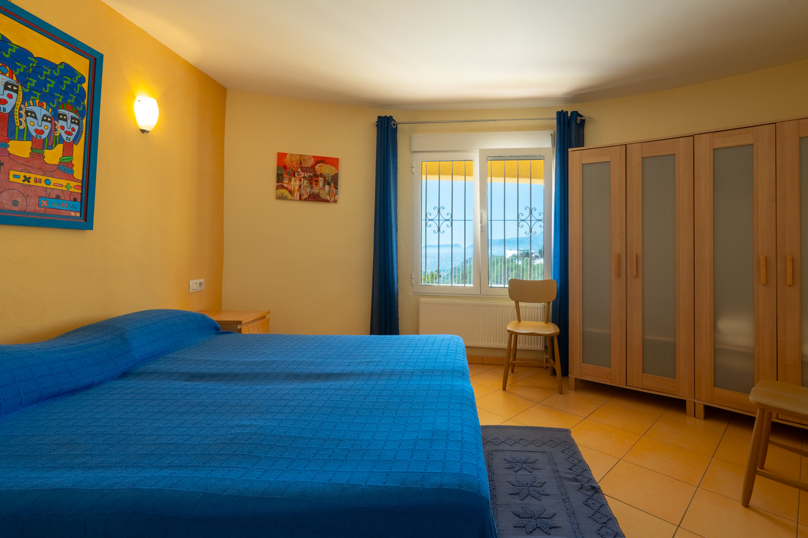Lichte slaapkamer met tweepersoonsbed, okergele muren, houten kledingkast met melkglas en raam met panoramisch uitzicht.