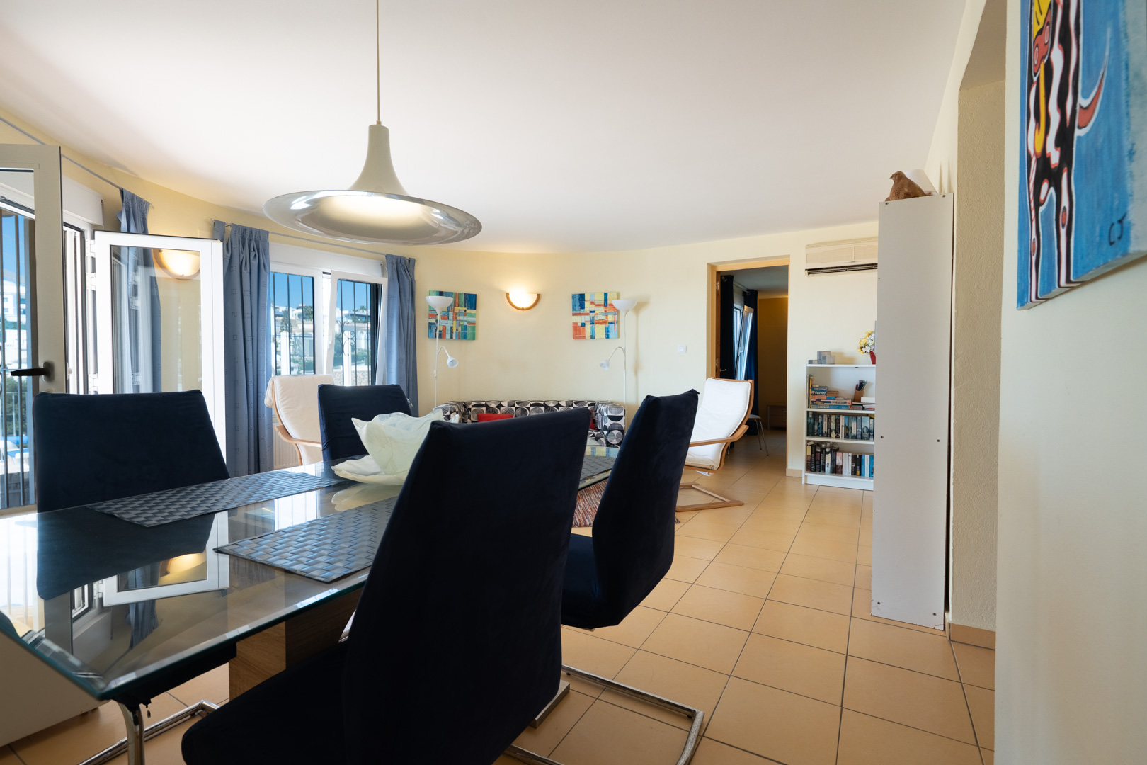 Lichtrijke eethoek met een glazen tafel en blauwe fluwelen stoelen, gelegen in een open woonruimte met keramische tegels en airconditioning.