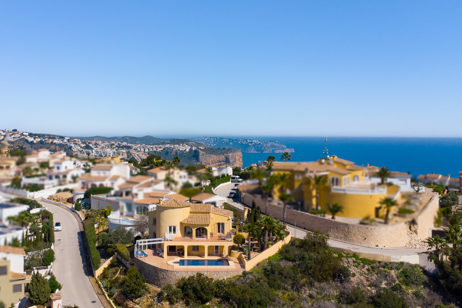 Luchtfoto van de gele villa met privézwembad en diverse terrassen, met panoramisch uitzicht over de Middellandse Zee en de Cap d’Or in Moraira.