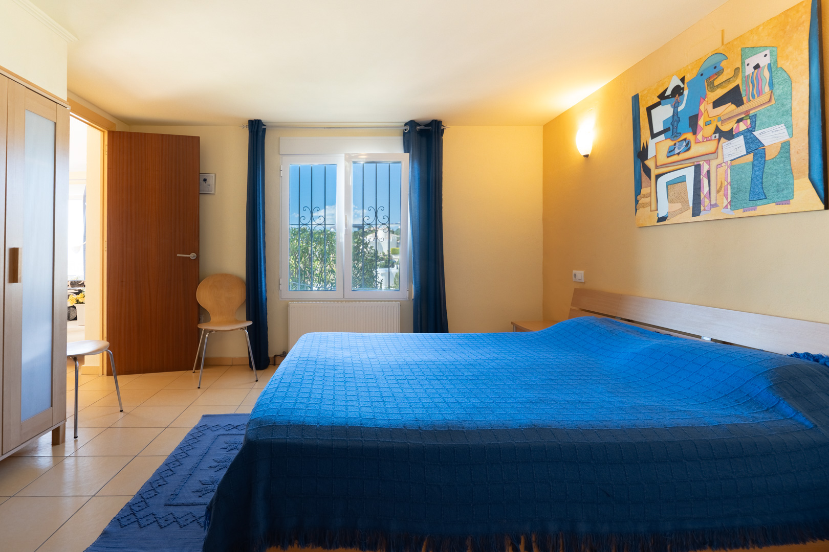 Slaapkamer met tweepersoonsbed, blauw textiel, houten kast en tegelvloer. Een raam met beveiligingstralies zorgt voor natuurlijke lichtinval.
