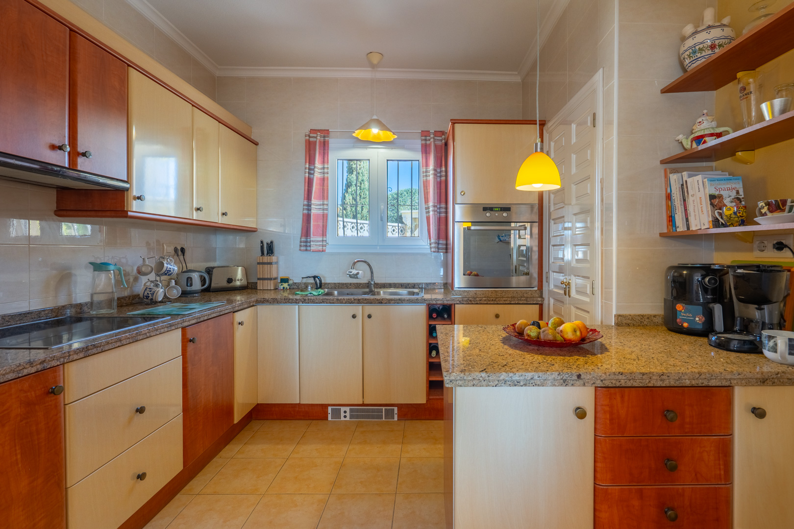 Volledig uitgeruste keuken met granieten werkbladen, tweekleurige kasten in hout en crème, en inbouwapparatuur. Voorzien van open schappen en natuurlijk daglicht via het raam.