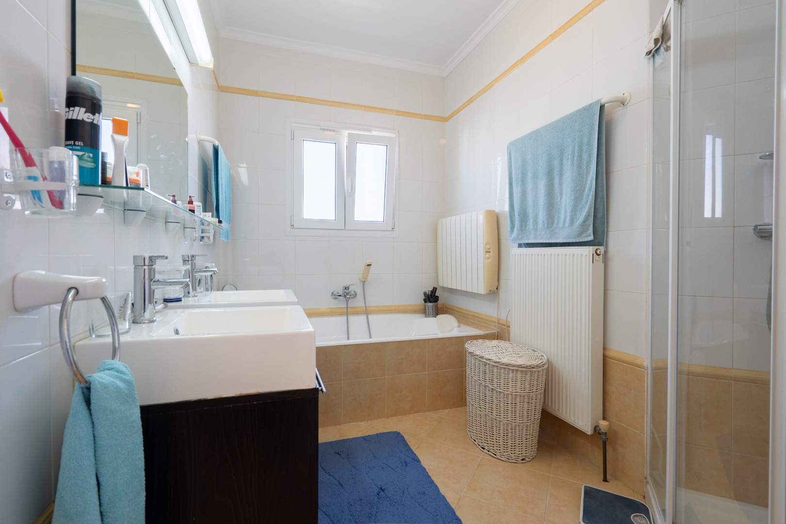Volledige badkamer met dubbele wastafel, ingebouwd ligbad, aparte douchecabine en witte wandtegels met een decoratieve gele bies.
