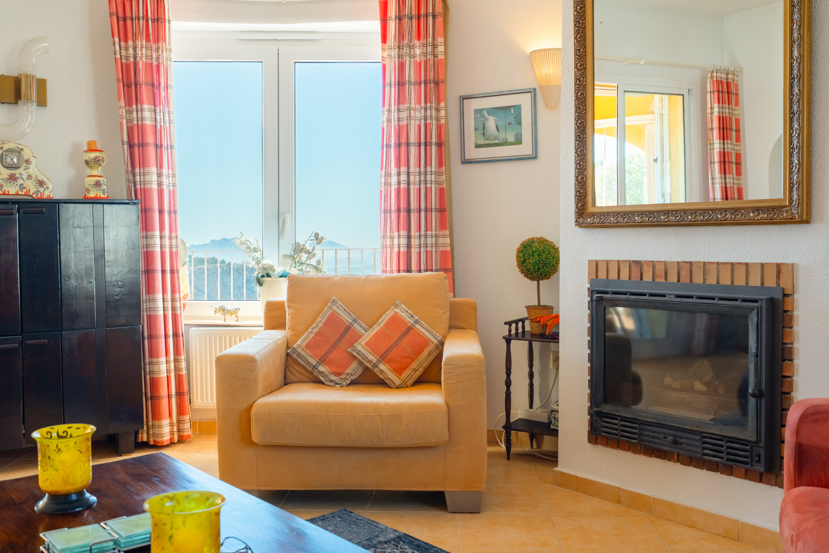 Woonkamer met ingebouwde open haard, een comfortabele fauteuil en een raam met uitzicht op de bergen in het binnenland en een grote spiegel.