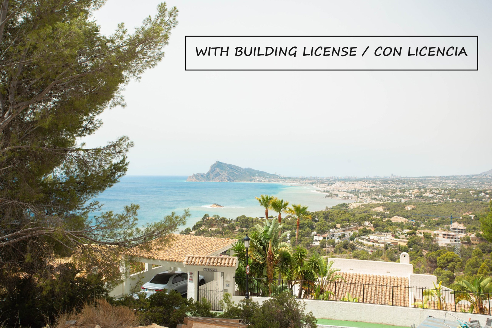 Panoramisch uitzicht vanaf het perceel over de baai van Altea en de skyline van Benidorm, omringd door mediterrane pijnbomen en lokale vegetatie.