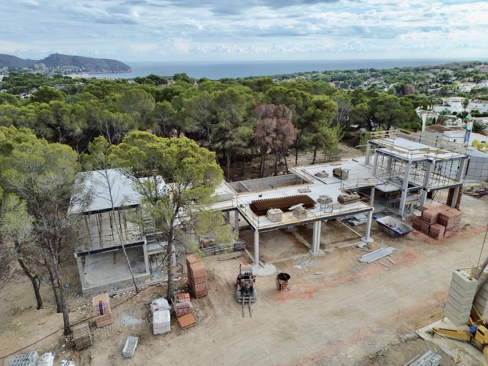 Luchtfoto van de in aanbouw zijnde villa, met de betonnen constructie en bouwmaterialen. Op de achtergrond zijn de Middellandse Zee en het bosrijke landschap zichtbaar.