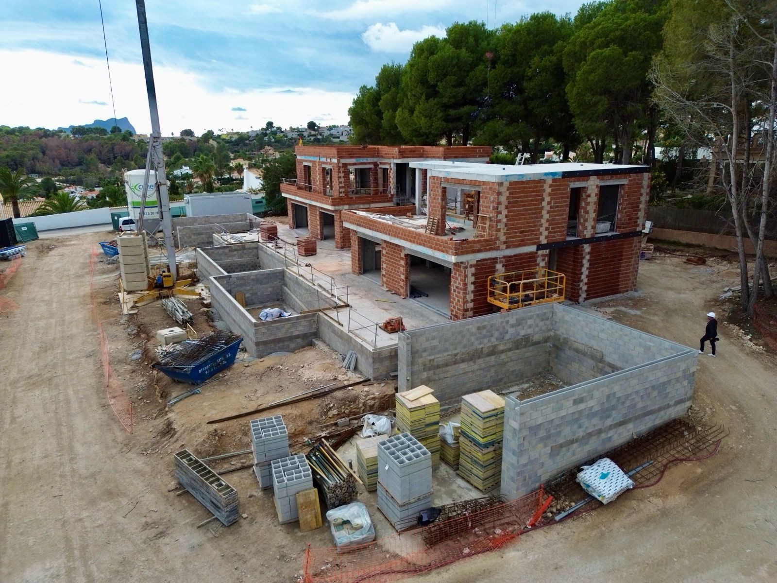 Luchtfoto van de villabouwplaats, met meerdere bakstenen structuren in aanbouw, uitgegraven betonnen funderingen en bouwmaterialen. Een kraan is zichtbaar, met verre groene heuvels.