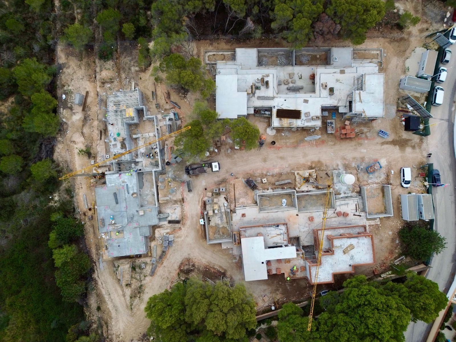 Luchtfoto van een villacomplex in aanbouw, met funderingen, betonplaten en twee kranen. Bouwmaterialen en voertuigen zijn zichtbaar op het terrein.