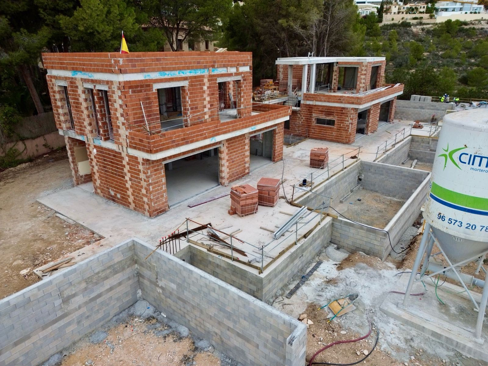 Luchtfoto van een villaproject in aanbouw, met het metselwerk van twee hoofdstructuren en betonblokfunderingen, gelegen op een groene heuvel.