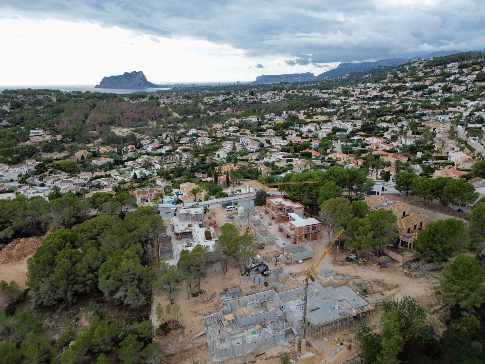 Luchtfoto van villa's in aanbouw in een woonwijk van Moraira, met zichtbare kranen en funderingen, omlijst door de zee en de Peñón de Ifach in de verte.