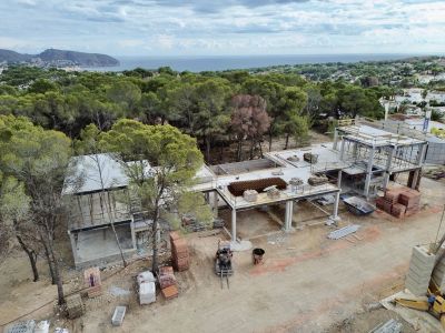 Luchtfoto van de in aanbouw zijnde villa, met de betonnen constructie en bouwmaterialen. Op de achtergrond zijn de Middellandse Zee en het bosrijke landschap zichtbaar.
