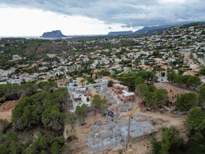 Luchtfoto van villa's in aanbouw in een woonwijk van Moraira, met zichtbare kranen en funderingen, omlijst door de zee en de Peñón de Ifach in de verte.