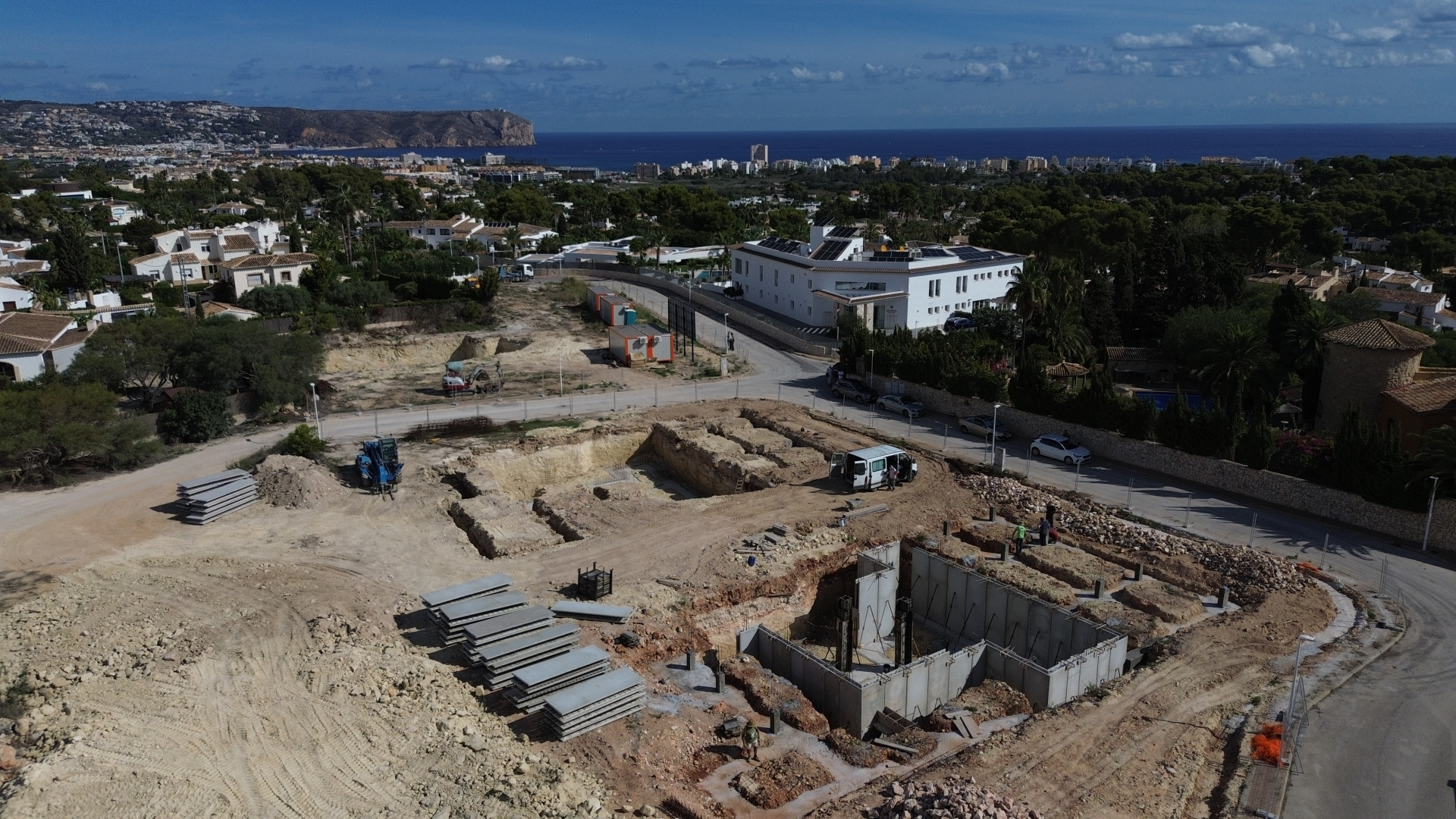 Luchtfoto van de bouwplaats met betonnen funderingen en grondwerken. Op de achtergrond zijn Jávea, de Middellandse Zee en Kaap San Antonio zichtbaar.