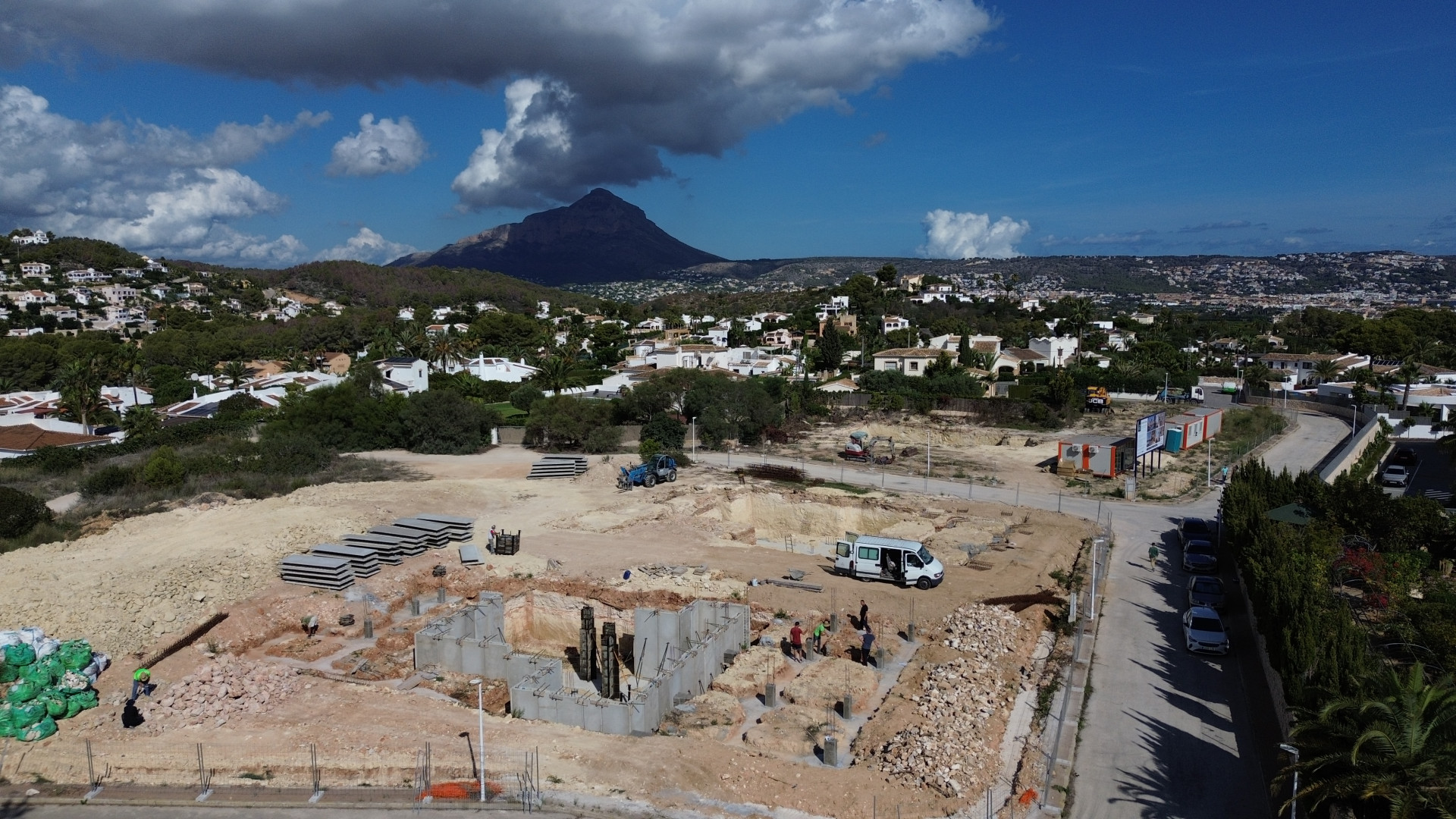 Luchtfoto van het bouwperceel in Jávea, met de fundering van de villa en bouwmaterialen. Op de achtergrond zijn woningen en de Montgó-berg zichtbaar.