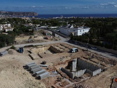 Luchtfoto van de bouwplaats met betonnen funderingen en grondwerken. Op de achtergrond zijn Jávea, de Middellandse Zee en Kaap San Antonio zichtbaar.