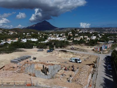 Luchtfoto van het bouwperceel in Jávea, met de fundering van de villa en bouwmaterialen. Op de achtergrond zijn woningen en de Montgó-berg zichtbaar.