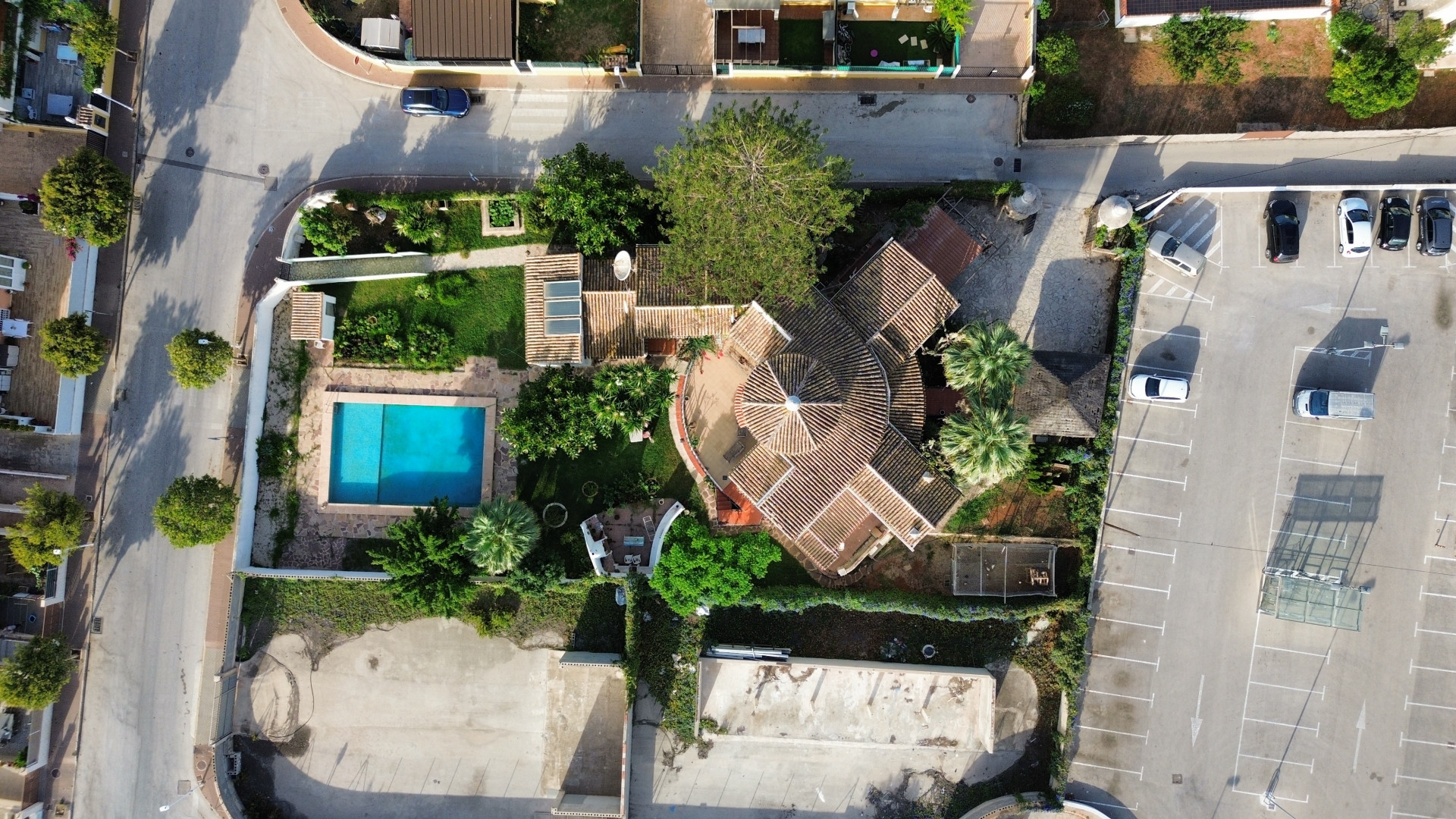 Luchtfoto van villa met zwembad en pannendak, omgeven door groen en aangrenzende parkeerplaats.