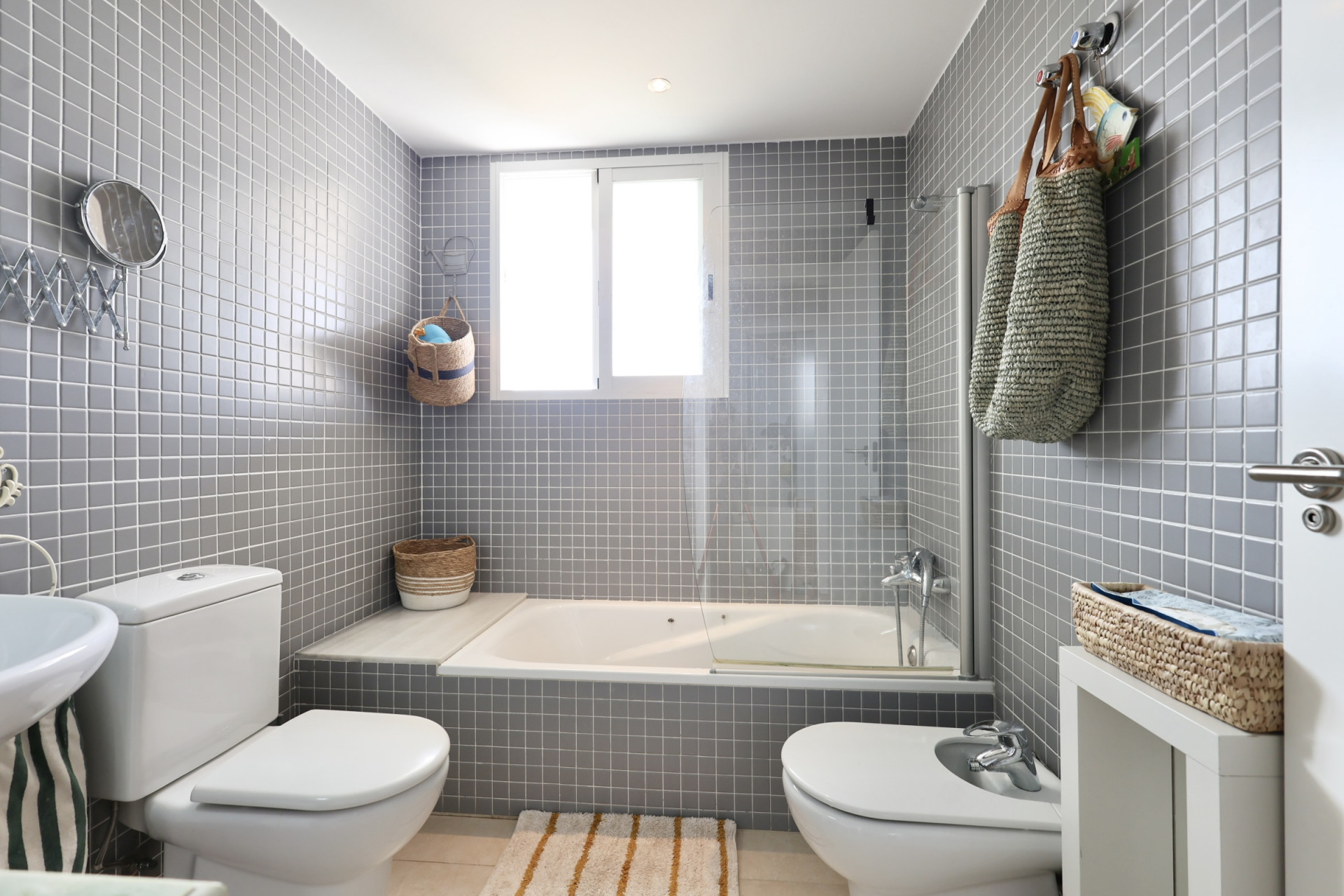 Badkamer met grijze tegelwanden, een ligbad met glazen scherm en decoratieve elementen van wicker.