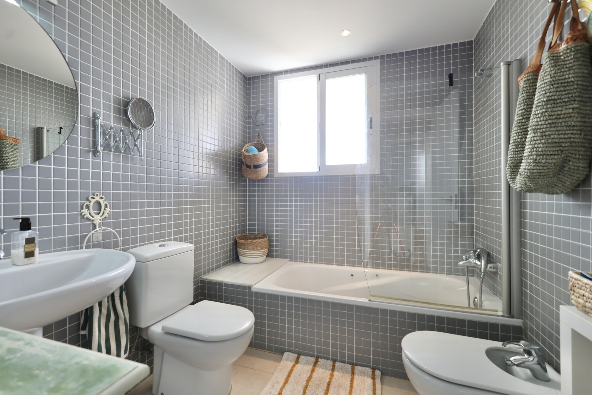 Badkamer met grijze vierkante tegels, wastafel op voetstuk, toilet en bidet. Een wit bad staat onder een raam.