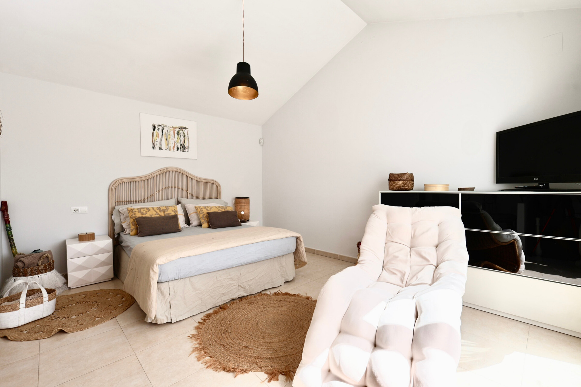 Lichte slaapkamer met rotan bed, jute vloerkleed en fauteuil naast een zwart tv-meubel.