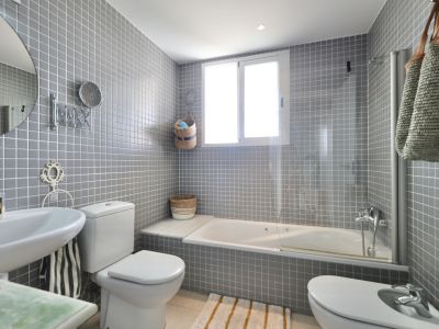 Badkamer met grijze vierkante tegels, wastafel op voetstuk, toilet en bidet. Een wit bad staat onder een raam.