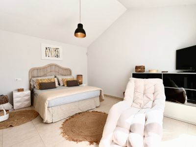 Lichte slaapkamer met rotan bed, jute vloerkleed en fauteuil naast een zwart tv-meubel.